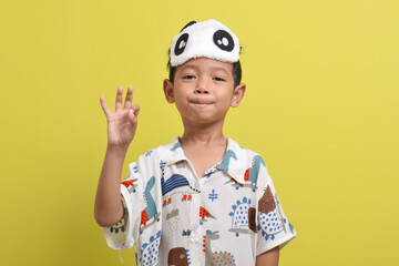Portrait of Asian child wearing pajamas and sleeping mask isolated on yellow background. Child wants to sleep. Healthy sleep concept. Copy space