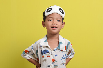 Portrait of Asian child wearing pajamas and sleeping mask isolated on yellow background. Child wants to sleep. Healthy sleep concept. Copy space