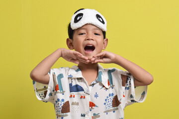 Portrait of Asian child wearing pajamas and sleeping mask isolated on yellow background. Child wants to sleep. Healthy sleep concept. Copy space