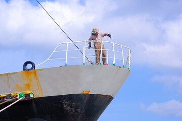 Asian blue collar worker is painting balustrade on the bow of the old oil tanker ship at harbor...