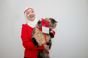Woman in white hijab wearing red sweater. celebrating Indonesia's independence day. holding a tabby cat and holding a flag. isolation on white background.