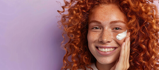 Fototapeta premium Portrait of a beautiful young woman with red curly hair smiling and applying cream to her face against a purple background, shown in closeup