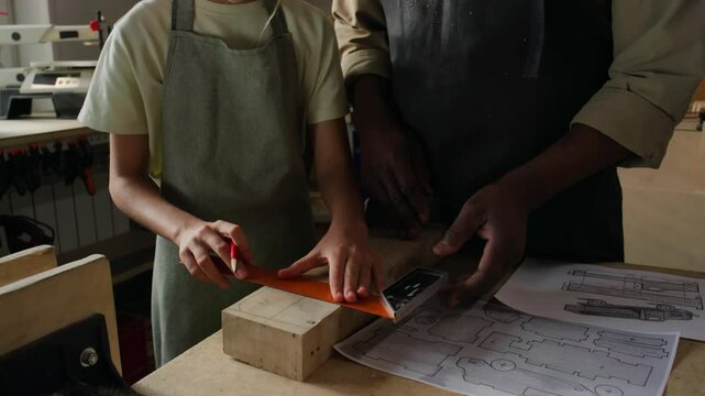 Close up view of hands of little girl and woodworking teacher drawing line on plank with pencil and ruler at workbench in carpentry workshop