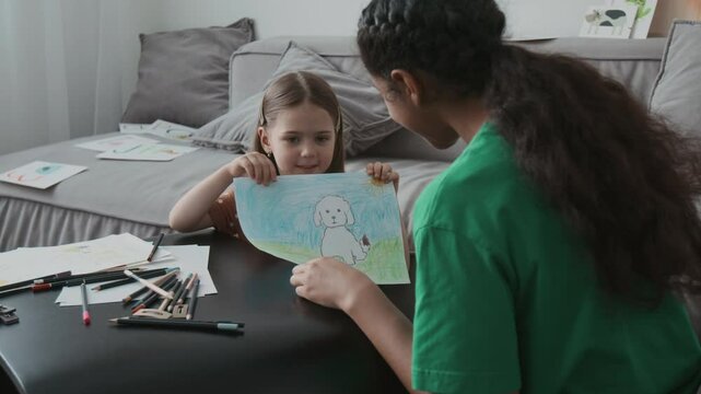 Slowmo shot of pretty Caucasian little girl giving her cute drawing of dog to African American teenage babysitter while staying at home together at leisure time