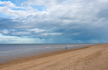 Two People Walk Along Sandy Beach With Overcast Sky and Rain Showers in Distance