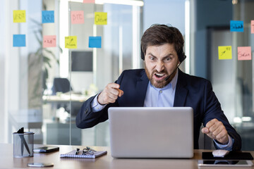 An angry businessman in a suit, shouting at his laptop while sitting at his desk in a modern office. The stress and frustration are evident as he clenches his fist and raises his voice.