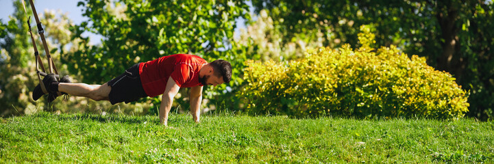 Fototapeta premium Focused and determined athlete, muscular strong man in sportswear doing trx workout in greenery park, standing in plank position with traps. Concept of sport, body and health care, active lifestyle