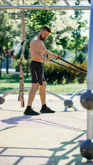 Obraz premium Male fitness enthusiast working out shirtless with resistance bands in park, surrounded by greenery and exercise equipment. Concept of sport, body and health care, active lifestyle, endurance