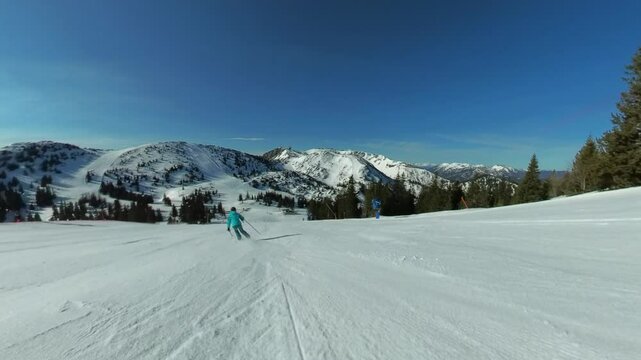 Following shot of a woman dressed in blue winter gear skiing fast on a ski slope with snow-covered mountains landscape