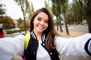 Portrait of adorable optimistic girl with brunette hairdo wear uniform rucksack making selfie smiling on green city street outdoors