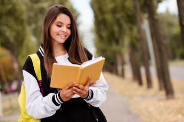 Fototapeta premium Portrait of clever nerdy girl with brunette hairdo wear uniform backpack read interesting story in book on green city street outdoors