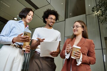 Three colleagues, engage in conversation while holding coffee and documents in an office lobby.