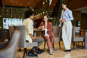 Three diverse colleagues chat over coffee during a break in a modern office setting.