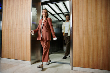 A woman with a prosthetic leg exits an elevator and smiles warmly near colleague who is stepping out.