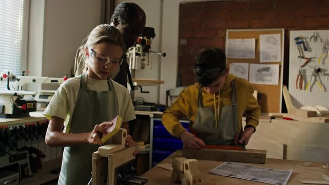 Two Caucasian children sanding and measuring wood pieces under supervision of Black male instructor in workshop with various tools and equipment