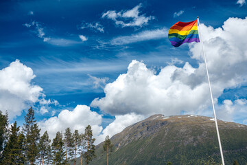 A pride flag hanging high on a flag pole with dramatic blue and cloudy skies and a mountain in the background