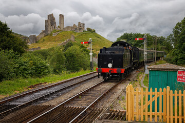 Fototapeta premium A steam train pulling into Corfe Castle Dorset UK with storm clouds in the sky and Corfe Castle ruins in the background