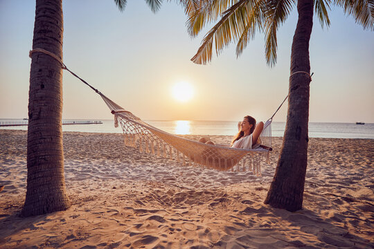 Young carefree inspired nomad woman with hands behind head relaxing in a hammock and enjoying working remotely at the laptop at seashore, dream job anywhere 