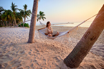 Young inspired contented freelancer girl nomad relaxing in a hammock and enjoying working remotely at the laptop at seashore, dream job anywhere 