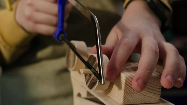 Close-up shot of hands of boy using hacksaw to cut wooden toy wheels during woodworking class
