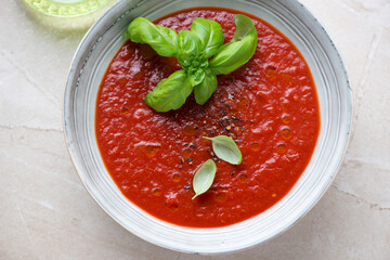 Bowl of tomato soup served with fresh green basil, horizontal shot on a beige stone surface, high angle view, middle close-up