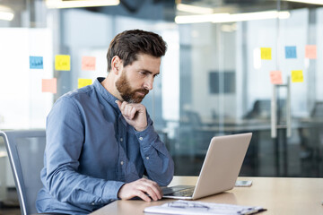 A serious and focused young man is sitting at a desk in the office and working on a laptop, looking...