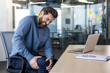 A young man sits in the office at the desk and holds his hands on his knee, grimaced and feels severe pain in his leg