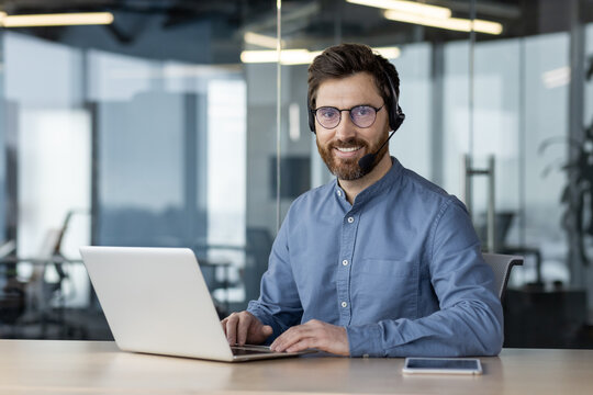 Portrait of a smiling young man wearing a headset sitting in the office at a desk with a laptop, looking confidently at the camera
