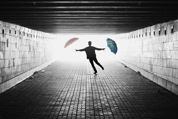 Silhouette of man with two umbrellas in a tunnel. Black and white image with light coming from behind. Person jumping towards the camera. Lens facing against the light.