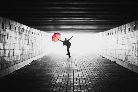 Silhouette of man with red umbrella in a tunnel. Black and white image with light coming from behind. Person jumping towards the camera. Lens facing against the light. - Powered by Adobe