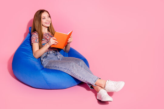 Full length photo of lovely teen sit bean bag read book look empty space dressed stylish print clothes isolated on pink color background