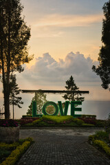 A captivating scene at Lake Toba, Sumatra, showcasing a 'LOVE' sign against a stunning sunrise backdrop. The serene lake, vibrant flowers, and lush greenery create a perfect romantic atmosphere.