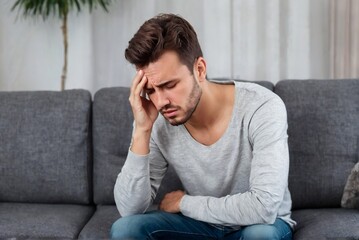 A man sits on a couch at home suffering from a headache or migraine with copy space.