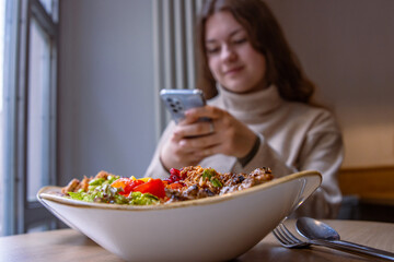 Young teenage girl surfing, scrolling, chatting, watching, texting, shopping online, checking her mobile phone while having lunch at the restaurant. Close up of healthy salad with beef Selective focus