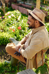 Bearded mature male gardener in workwear and hat looking forwards while sitting in front of...