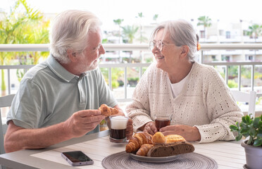 Carefree senior couple enjoying a morning breakfast together at a cozy outdoor balcony with croissant and coffee and tea, authentic serene retirement lifestyle