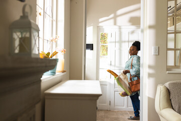 Happy senior african american woman opening door and holding shopping bag at home