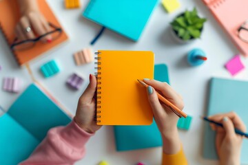 Close up of hands holding an open notebook with a pencil, surrounded by colorful stationery and other items.