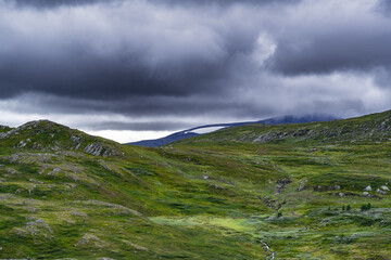 A sweeping view of rolling green hills and a distant mountain range, all under a dramatic sky with dark, swirling clouds in Norway