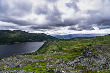 A panoramic view from a rocky hilltop overlooking a long, narrow lake in Norway. Lush green hills and valleys surround the lake, with a cloudy sky above