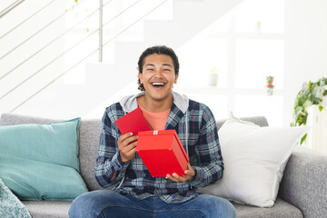 Young biracial man looks surprised during a video call as he opens a gift at home
