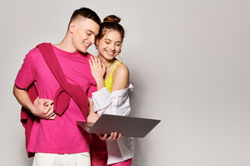 A stylish young couple stands with laptop side by side in a studio with a grey background.