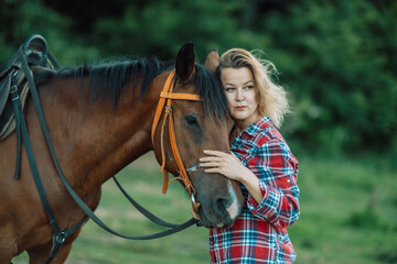 Obraz premium Happy blonde with horse in forest. Woman and a horse walking through the field during the day. Dressed in a plaid shirt and black leggings.