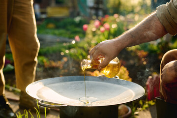 Hand of unrecognizable male farmer pouring olive oil on large round plate standing on iron barrel...
