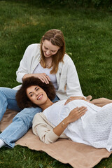 A pregnant woman rests on the lap of her partner while they relax on a blanket in a grassy park.