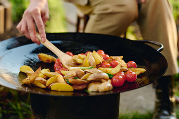 Hand of unrecognizable woman with wooden spatula mixing roasted chopped potatoes, red tomatoes and green leek on frying pan