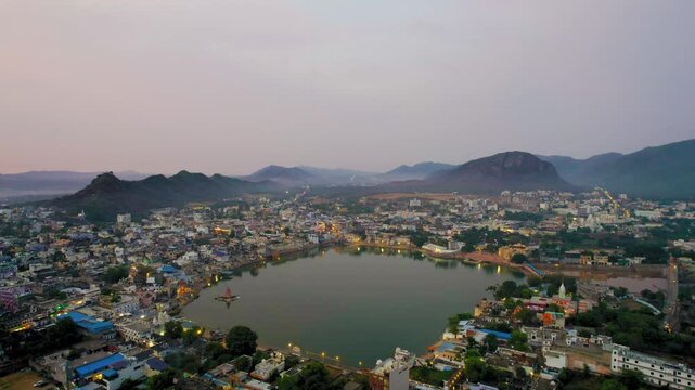 Aerial drone side flying shot over hindu holy city of pushkar with lake bhrama temple visible in city lights and aravalli hills covered in fog in distance showing cityscape in Rajasthan India