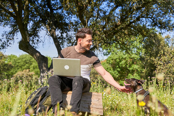 digital nomad, young Caucasian working with his laptop sitting in the forest, backpacking, petting his dog © Oscar