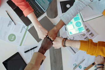 Close-up of diverse team members fist bumping over desk in office. Display of unity and teamwork with digital devices and documents spread out in background