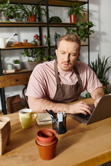 A man sits at a table in a plant shop, focused on his laptop, embodying the owner of a successful floral business.